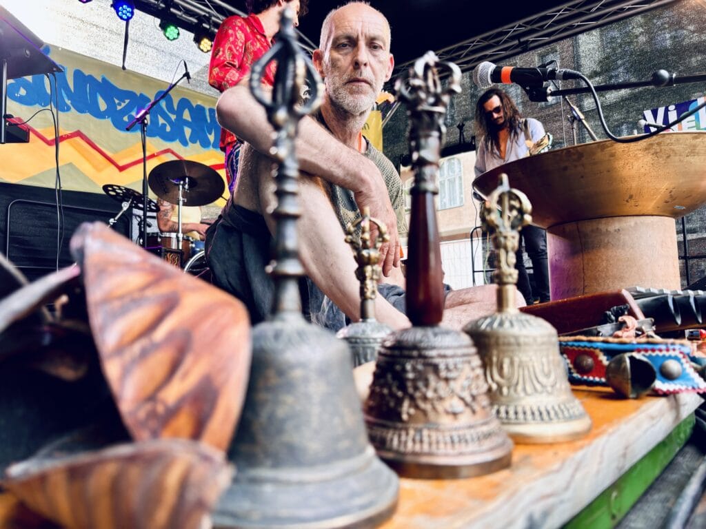 Ein Mann sitzt vor einem Tisch mit verschiedenen Klanginstrumenten, während im Hintergrund eine Band auf der Bühne spielt.