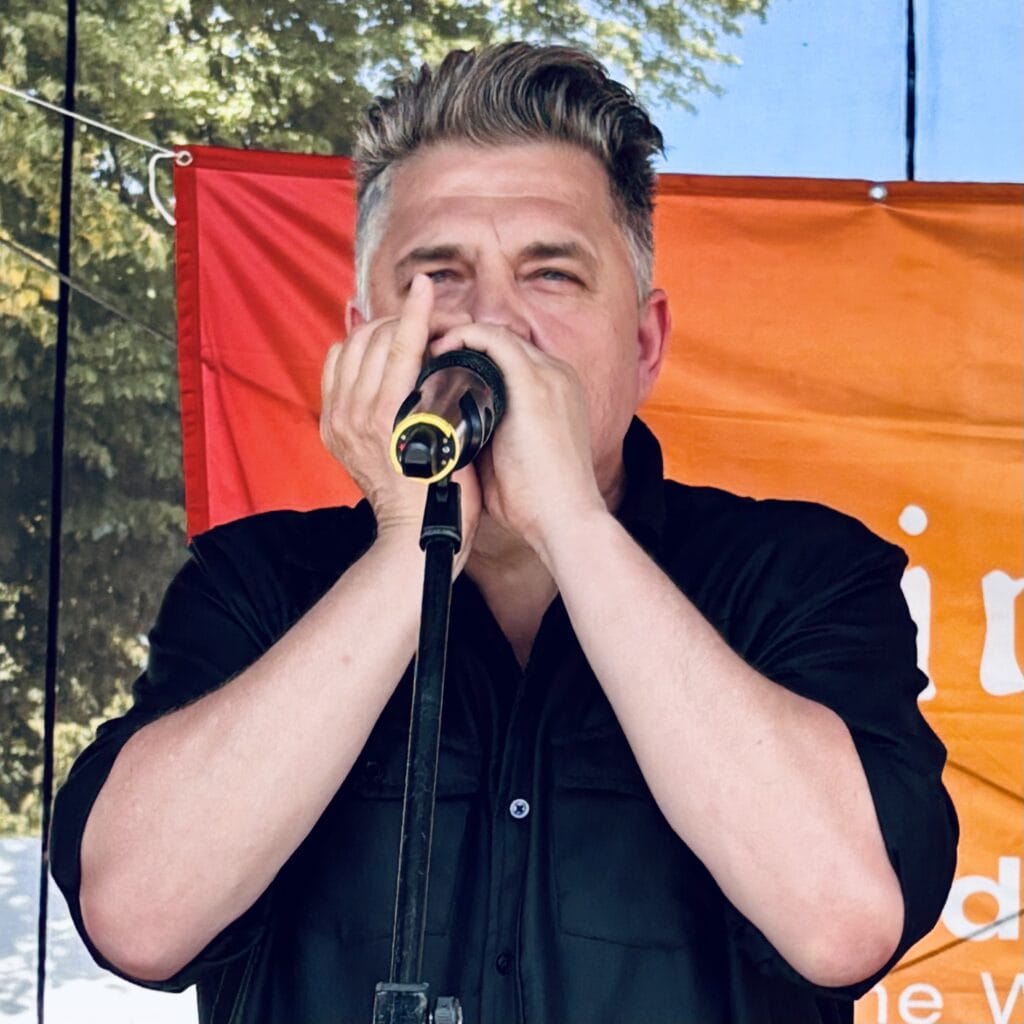 Ein Mann im schwarzen Hemd spielt Mundharmonika und singt in ein Mikrofon bei einer Veranstaltung.