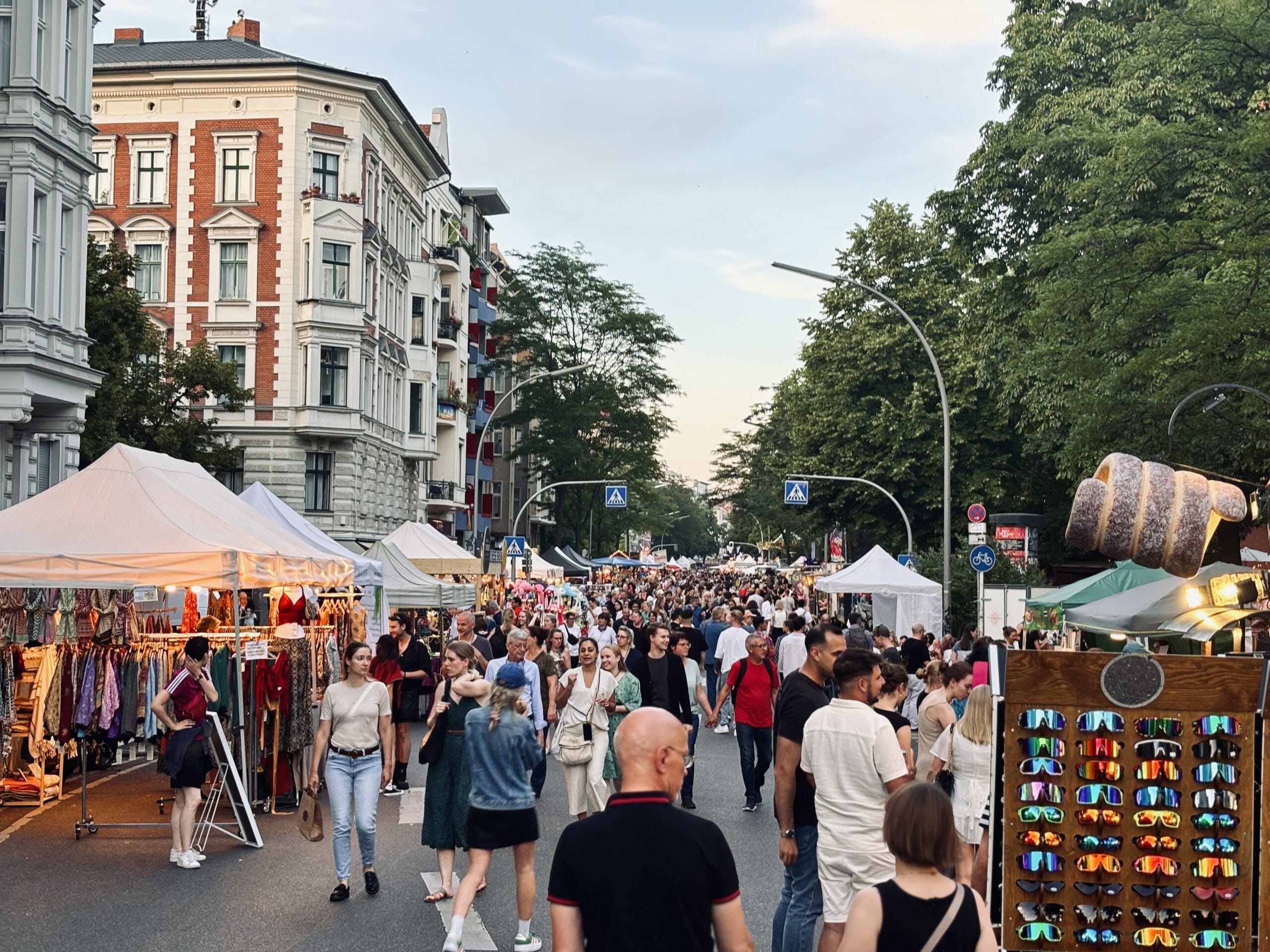 Eine Menschenmenge flaniert über eine Straße, flankiert von Verkaufsständen und alten Wohnhäusern, an einem sonnigen Tag in Berlin.