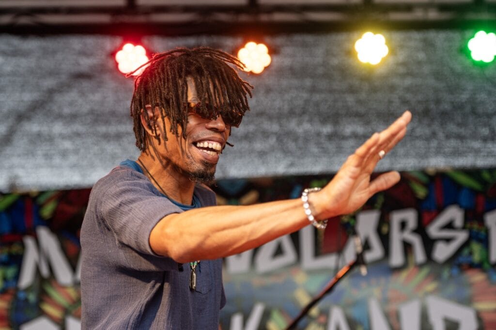 Ein Mann mit Dreadlocks und Sonnenbrille performt auf einer Bühne mit bunten Lichtern im Hintergrund.