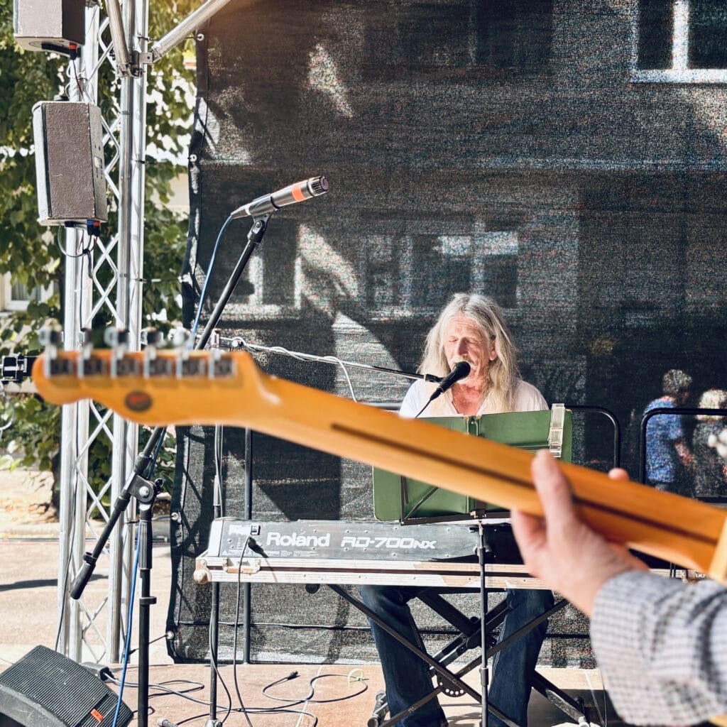 Ein Musiker mit langen Haaren spielt Keyboard und singt auf einer Bühne, während eine Gitarre im Vordergrund zu sehen ist.