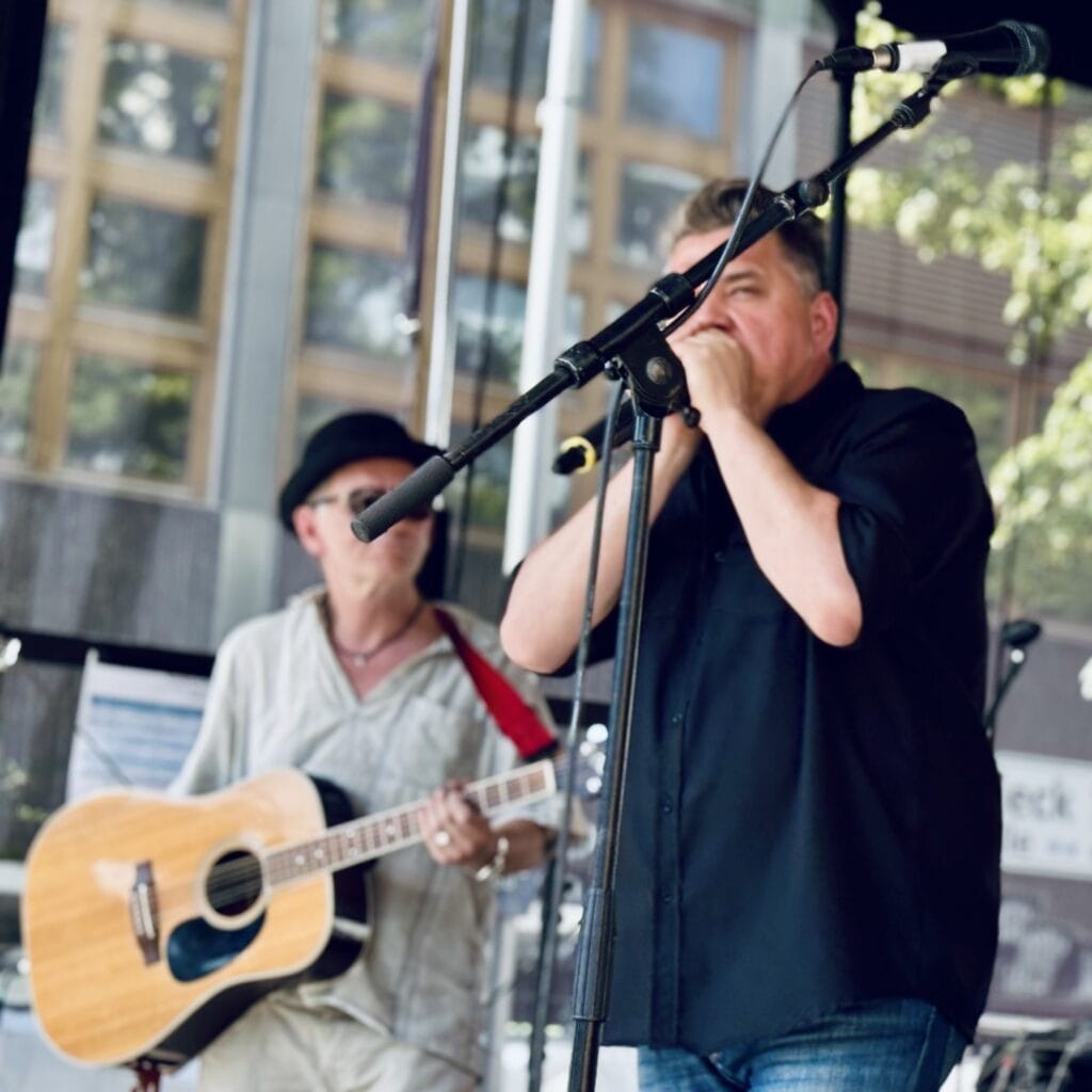 Ein Mann spielt Mundharmonika am Mikrofon, ein anderer spielt Gitarre auf einer Open-Air-Bühne.