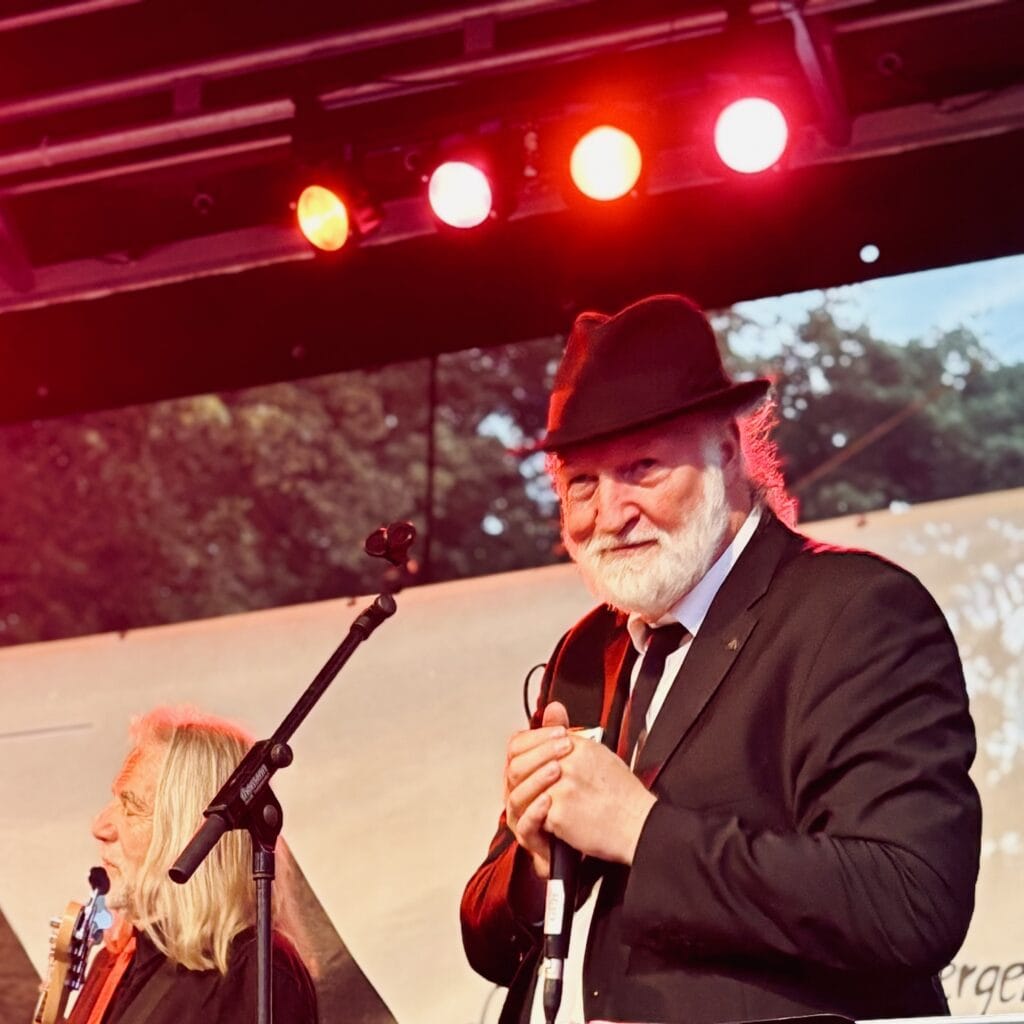 Ein älterer Musiker mit Hut steht auf der Bühne und singt in ein Mikrofon, Gitarrist im Hintergrund.