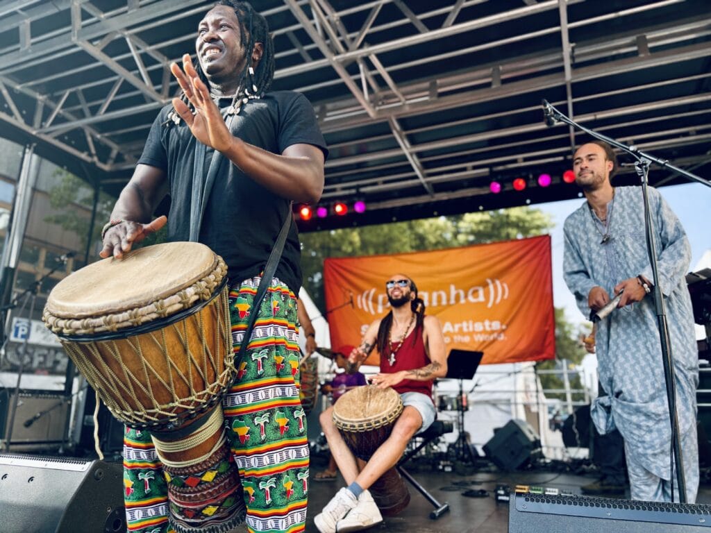 Ein Mann mit Dreadlocks und bunten Hosen spielt auf einer Trommel während eines Auftritts mit Band.