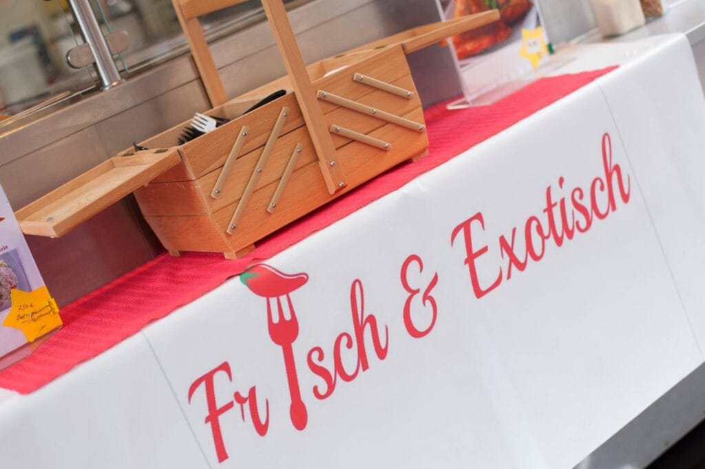Ein Stand mit der Aufschrift 'Frisch & Exotisch' mit einer Gabel und einer Chili-Schote als Logo. Eine Holzbox steht daneben.