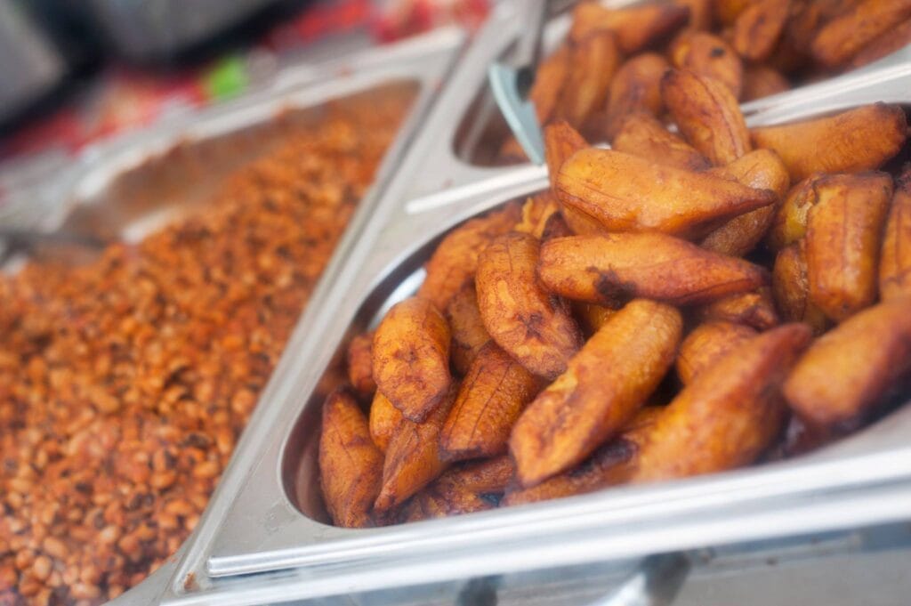 Auf einem Fest werden goldbraun gebratene Kochbananen in einer Metallschale präsentiert, daneben weitere Speisen.