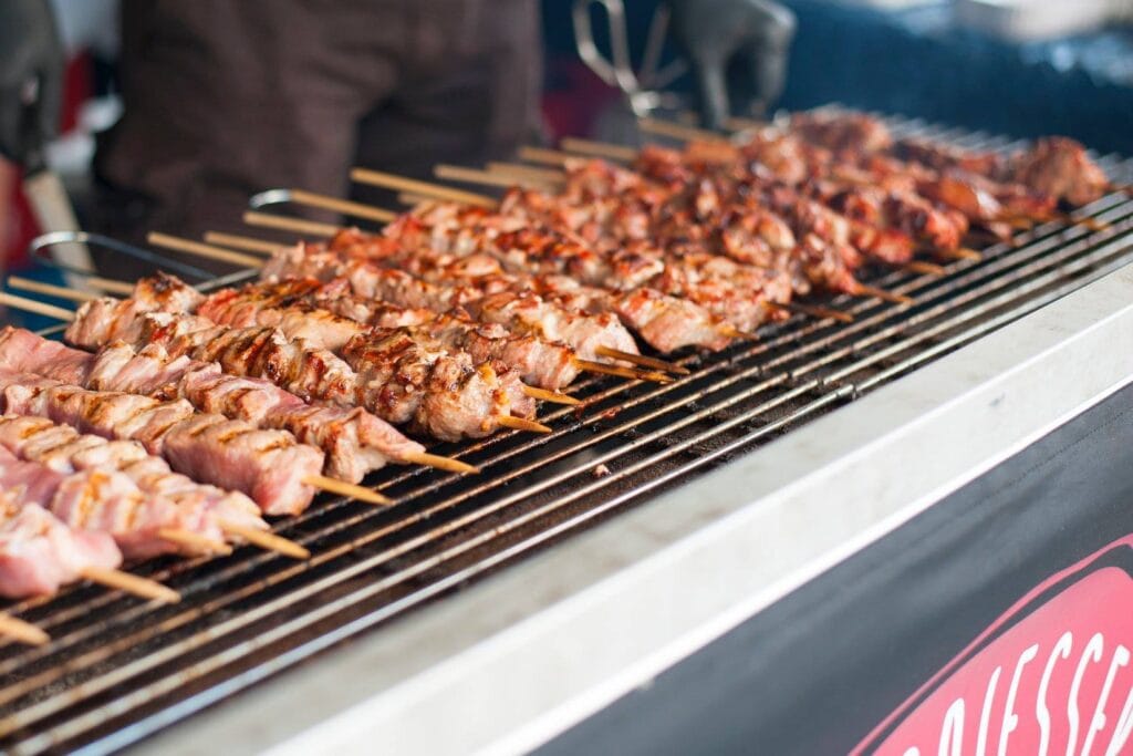 Auf dem Grill liegen viele Spieße mit Fleisch, die von einem Koch gewendet werden.