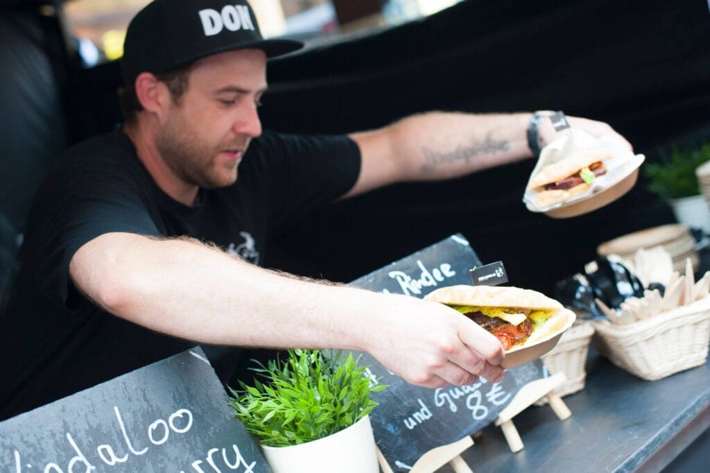 Ein Verkäufer reicht ein Sandwich über die Theke; auf Schiefertafeln stehen die Speisen.
