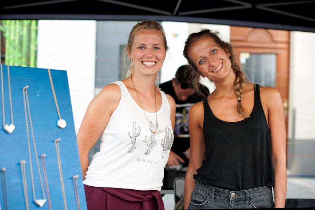 Zwei junge, lächelnde Frauen präsentieren ihren Schmuck auf einem Markt, der auf einem blauen Brett ausgestellt ist.