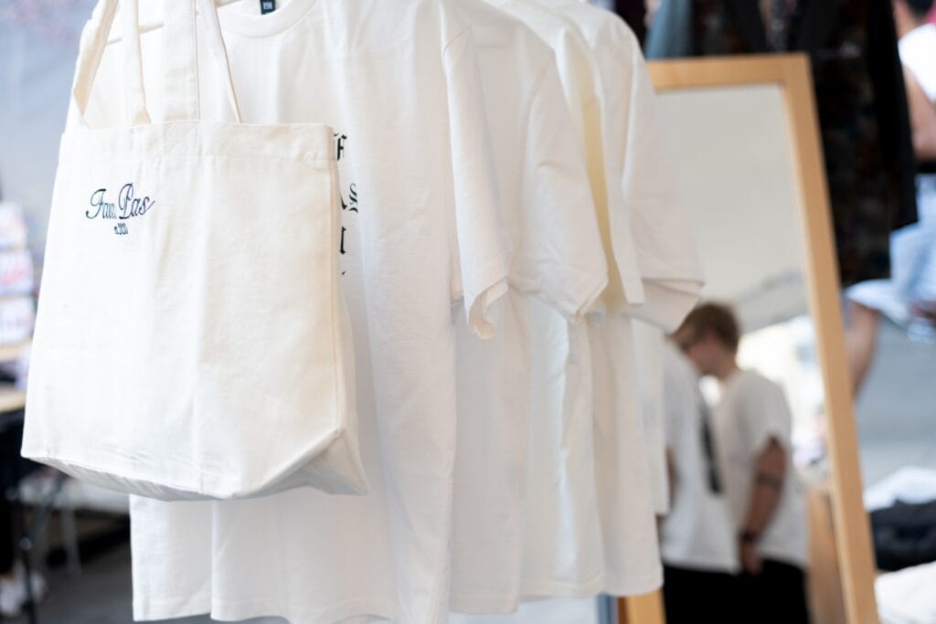 Aufgereihte, weiße T-Shirts hängen neben einem Stoffbeutel mit Aufdruck; eine Person spiegelt sich leicht im Hintergrund.