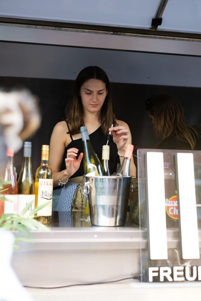 Eine junge Frau schenkt an einem Weinstand im Freien Wein ein, Weinflaschen und ein Eiskühler sind zu sehen.