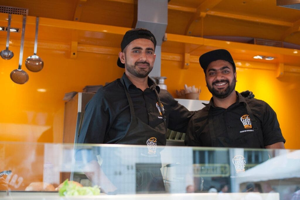 Zwei freundliche Verkäufer mit Schürzen von Liebes Beef stehen hinter der Theke und bereiten Burger zu.