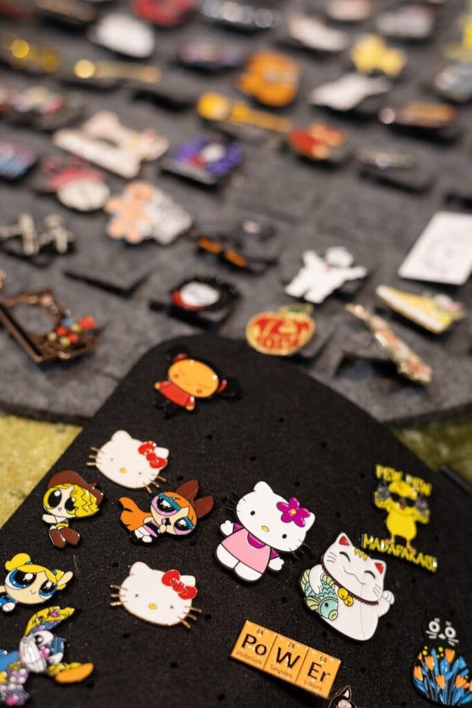 Eine vielfältige Auswahl an bunten Emaille-Pins, darunter Hello Kitty, Powerpuff Girls und andere Motive auf zwei grauen Filzunterlagen.