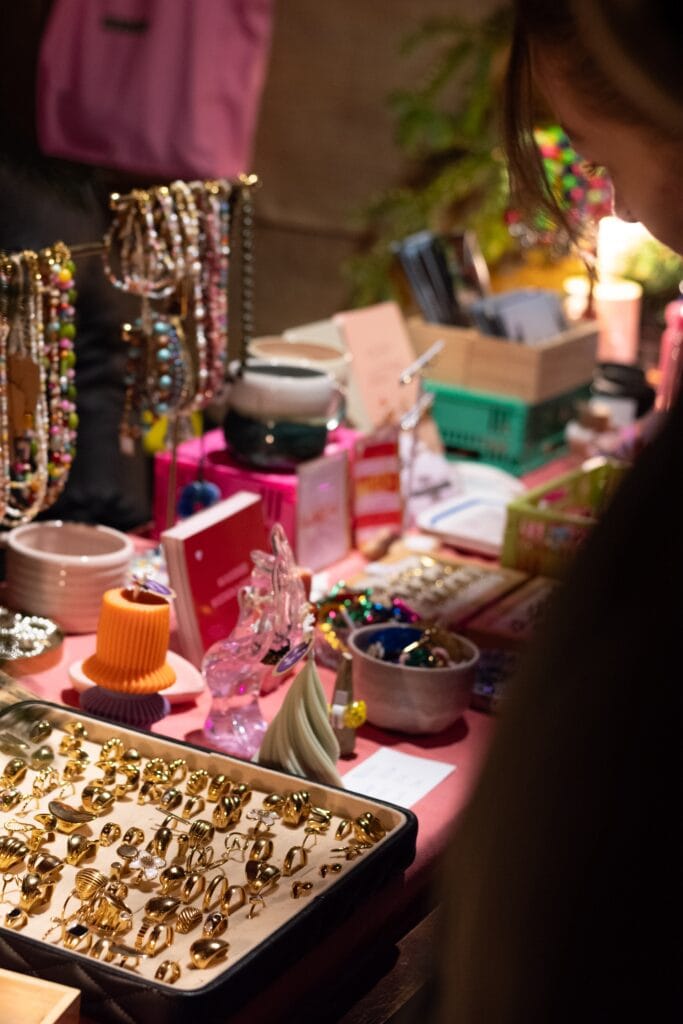 Auf einem festlichen Markt betrachtet eine Person goldfarbene Schmuckstücke, die auf einem Tisch mit weiterem Schmuck angeboten werden.