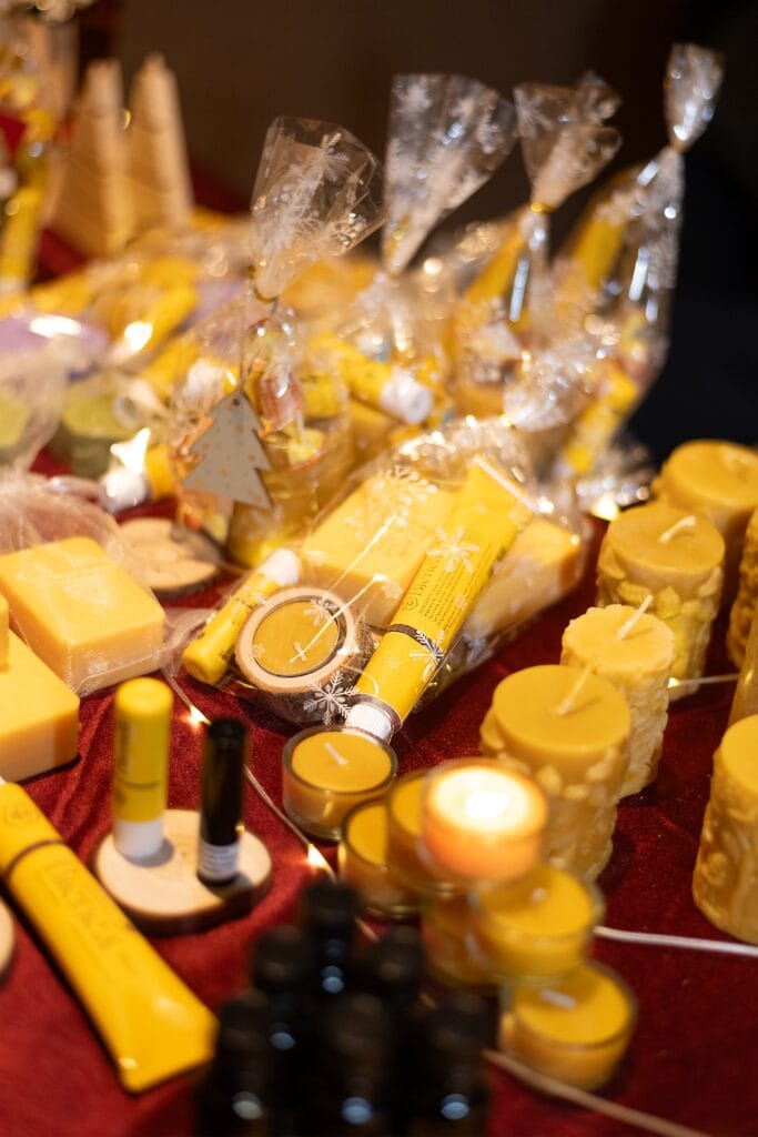 Auf einem roten Tuch liegen verschiedene handgemachte Produkte aus Bienenwachs, darunter Kerzen, Kosmetika und Seifen, ein Teelicht brennt hell.