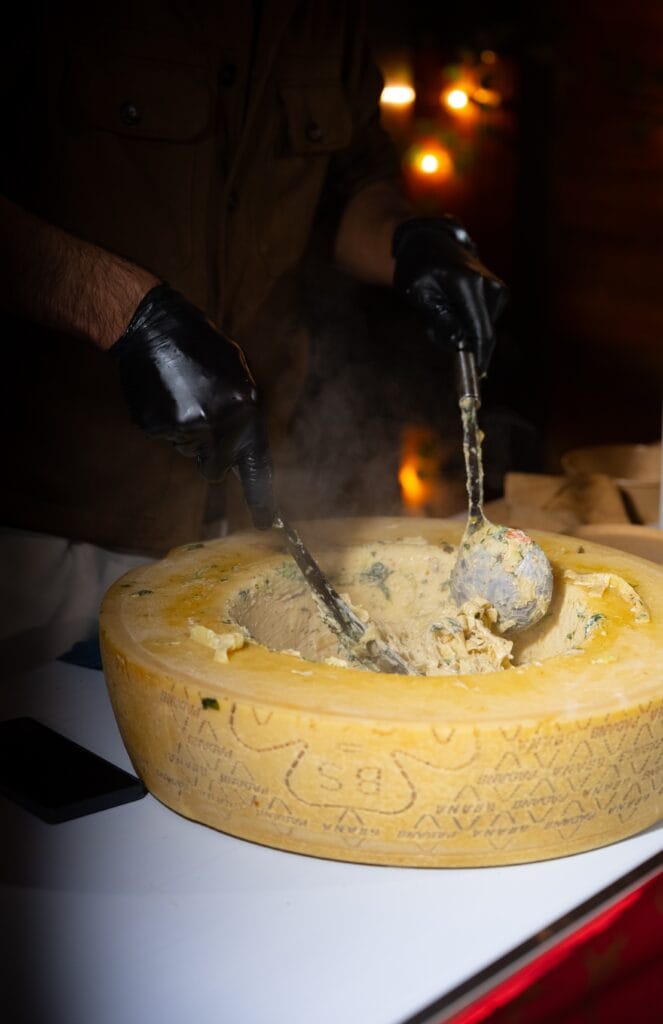 Ein Koch mit schwarzen Handschuhen bereitet Pasta in einem großen Käselaib zu, Dampf steigt auf.