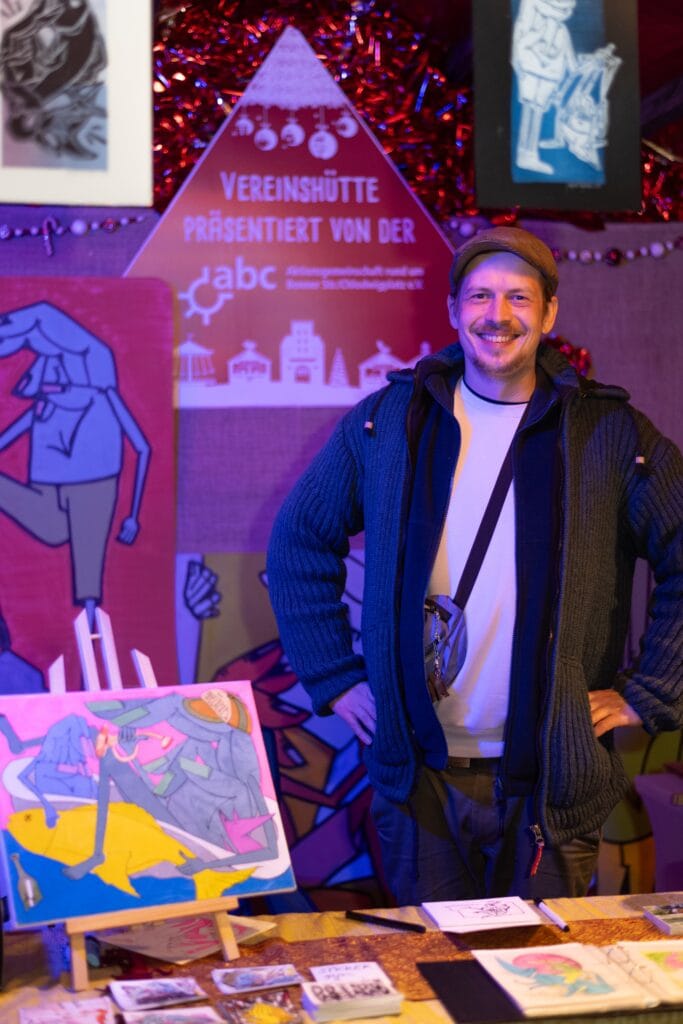 Ein lächelnder Mann mit Mütze steht an einem Stand mit Kunstwerken und Flyern auf dem Veedelsadvent.
