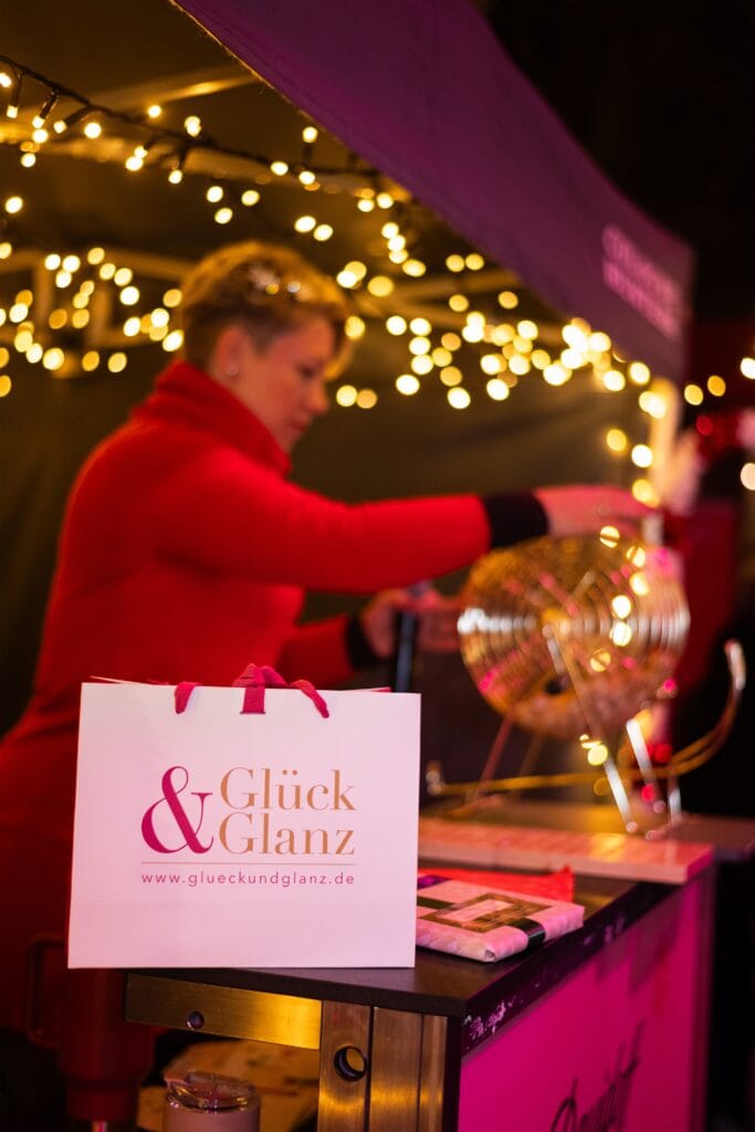 Eine Losverkäuferin kurbelt an einer Losbude, während eine Geschenktasche von "Glück & Glanz" im Vordergrund steht.