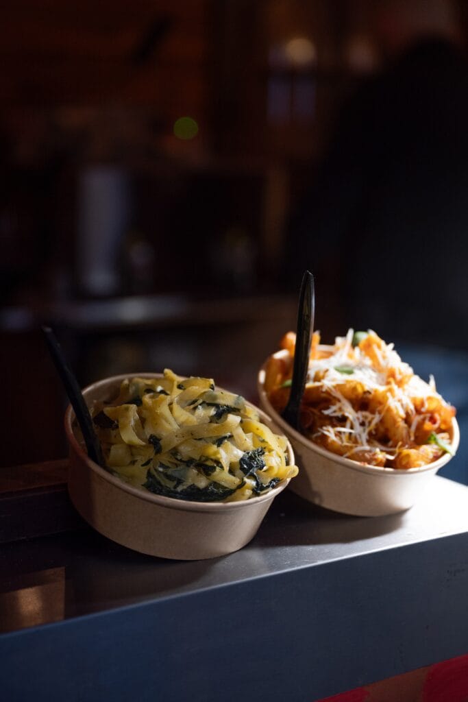 Zwei Pappbecher mit köstlich aussehender Pasta, einmal mit Spinat und einmal mit Käse überbacken, stehen auf einem Tresen.