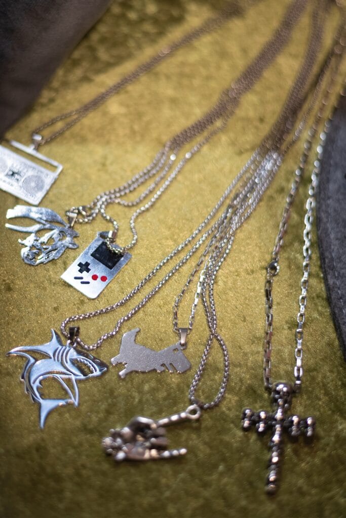 Mehrere Silberketten mit unterschiedlichen Anhängern liegen auf einem grünen Stoff, darunter ein Gamecontroller, ein Hai und ein Totenkopf-Kreuz.