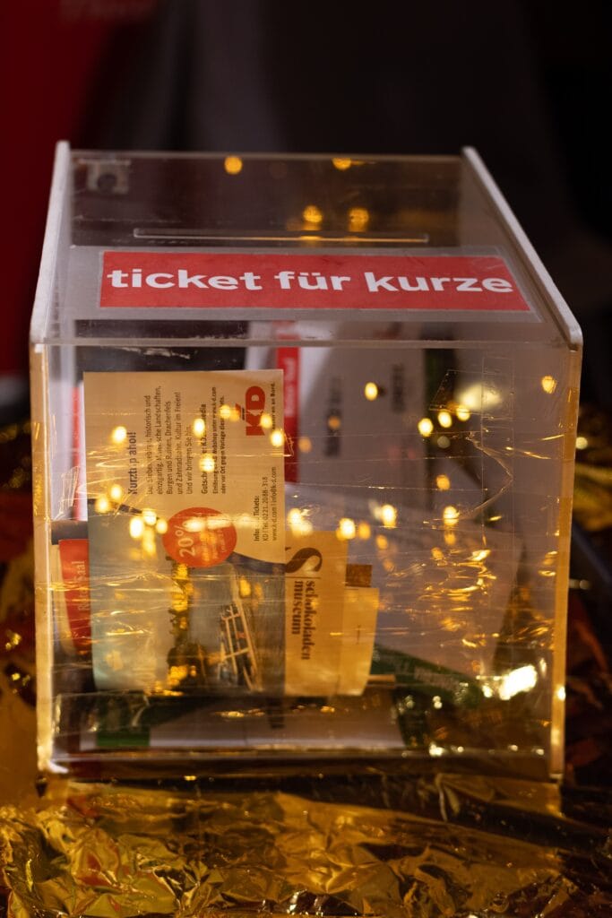 Eine durchsichtige Spendenbox gefüllt mit Flyern und Tickets, beschriftet mit 'Ticket für kurze', steht auf einer goldenen, glänzenden Oberfläche.