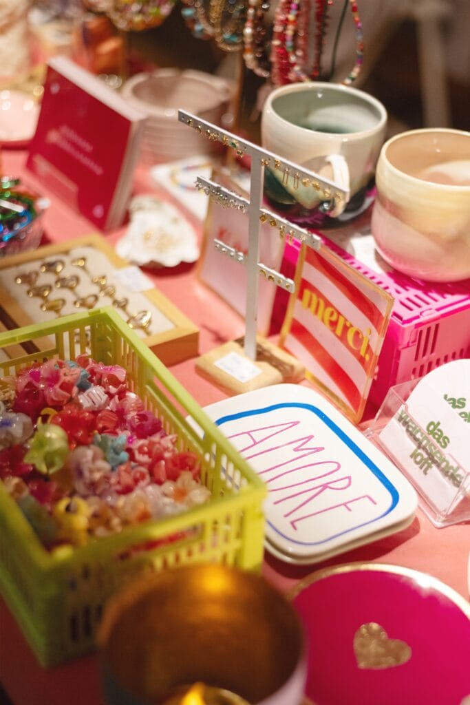 Ein bunter Verkaufsstand präsentiert handgefertigten Schmuck, Dekorationsobjekte und Accessoires auf einem Tisch mit rosa Oberfläche.