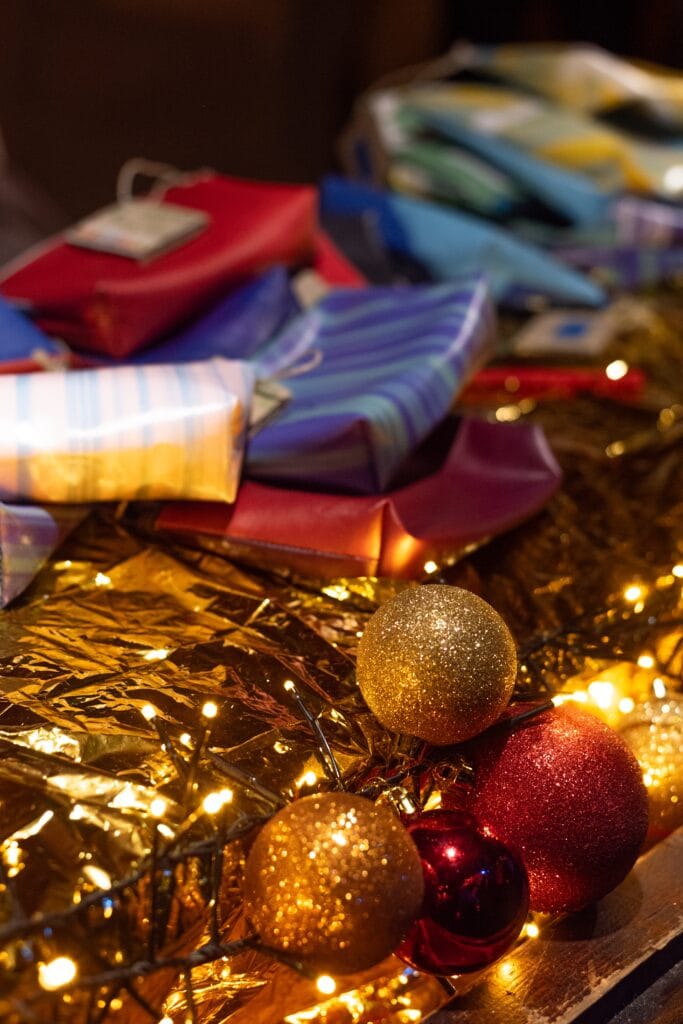 Funkelnde Weihnachtskugeln und Lichterketten liegen neben bunten, verpackten Geschenken auf goldenem Untergrund und erzeugen eine festliche Atmosphäre.
